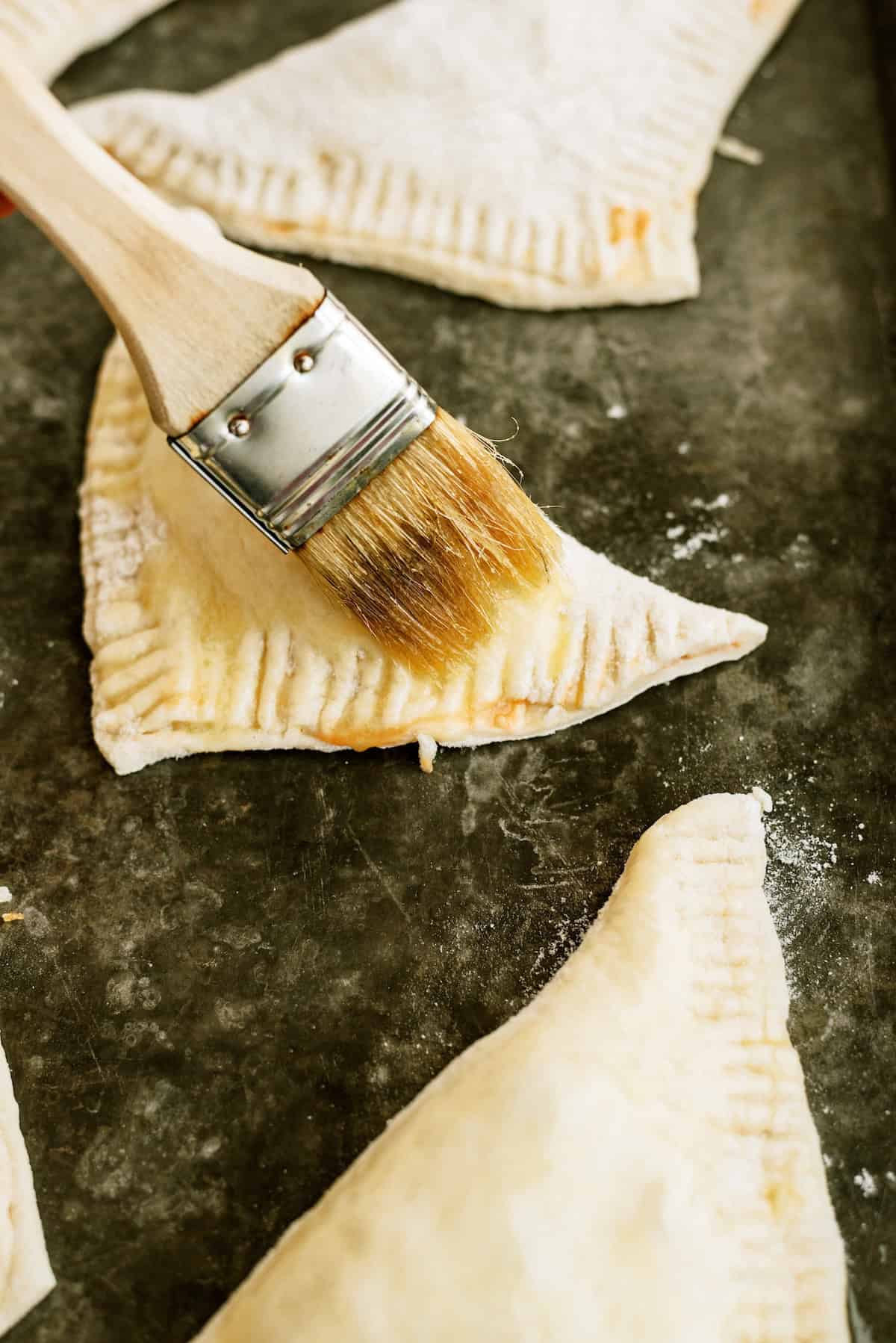 A pastry brush applies an egg wash to a triangular piece of unbaked pastry dough on a floured baking sheet.
