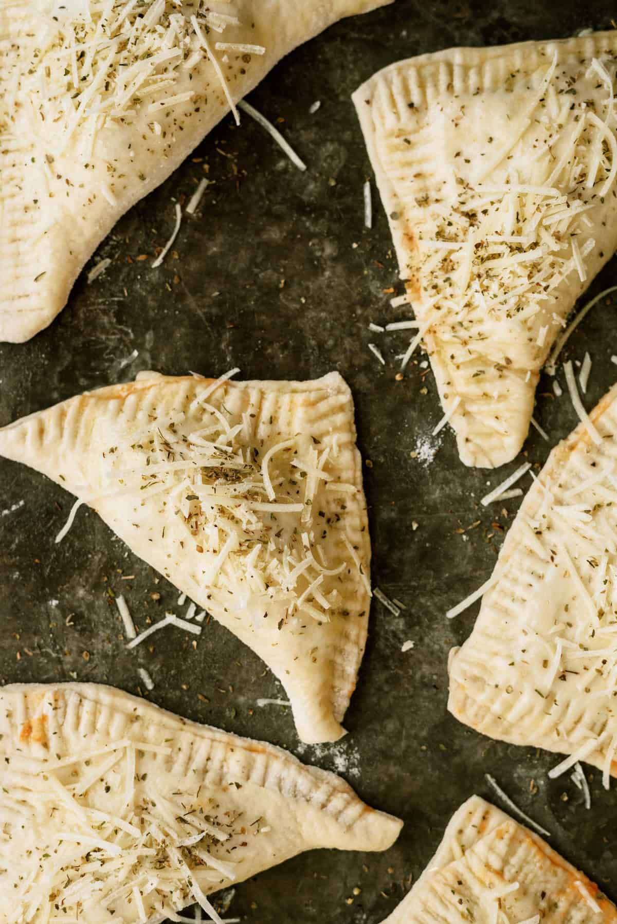 Unbaked triangular pastry turnovers are arranged on a dark baking sheet, topped with shredded cheese and herbs.