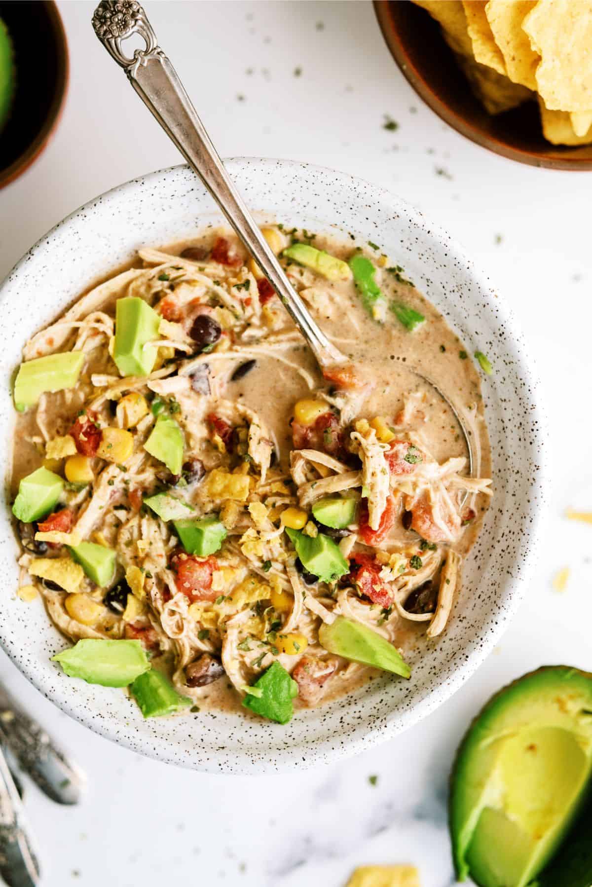 A bowl of creamy chicken soup with shredded chicken, black beans, diced tomatoes, chopped avocado, and herbs, with a spoon in the bowl and tortilla chips on the side.