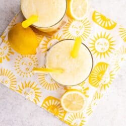 Two glasses of lemon drink with yellow straws sit on a sun-patterned napkin, next to a whole lemon and lemon slices, on a light-colored surface.