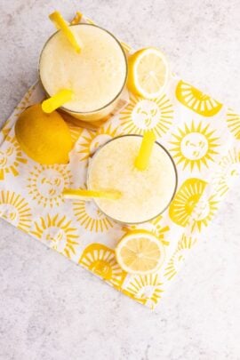 Two glasses of lemon drink with yellow straws sit on a sun-patterned napkin, next to a whole lemon and lemon slices, on a light-colored surface.