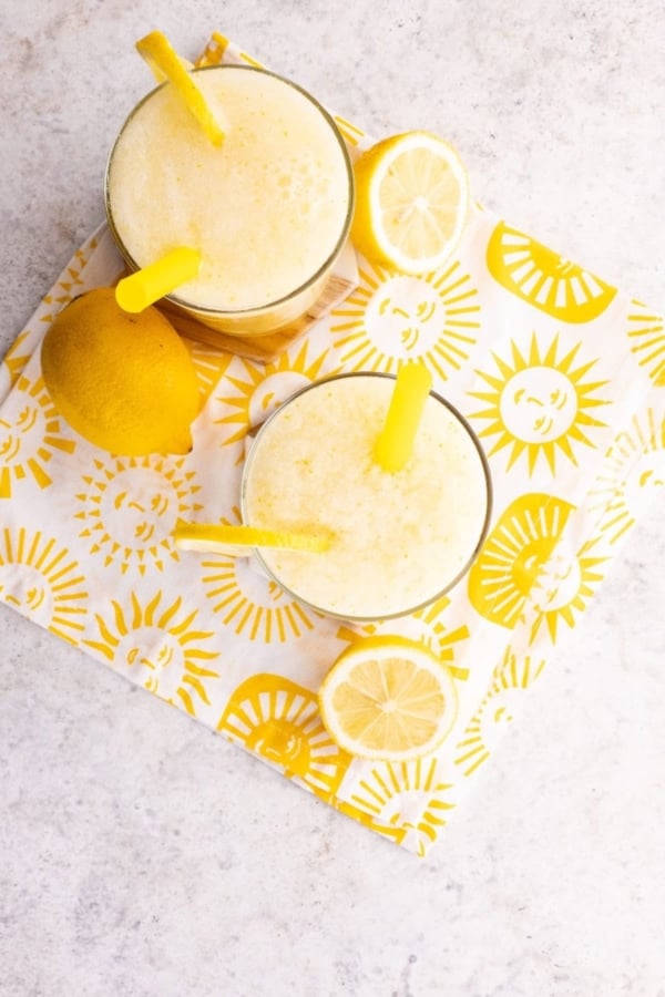Two glasses of lemon drink with yellow straws sit on a sun-patterned napkin, next to a whole lemon and lemon slices, on a light-colored surface.