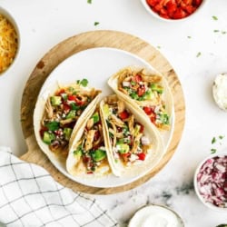 Three pork tacos on a white plate with toppings; surrounded by bowls of diced tomatoes, shredded cheese, sour cream, red onion, and a bowl of shredded chicken.