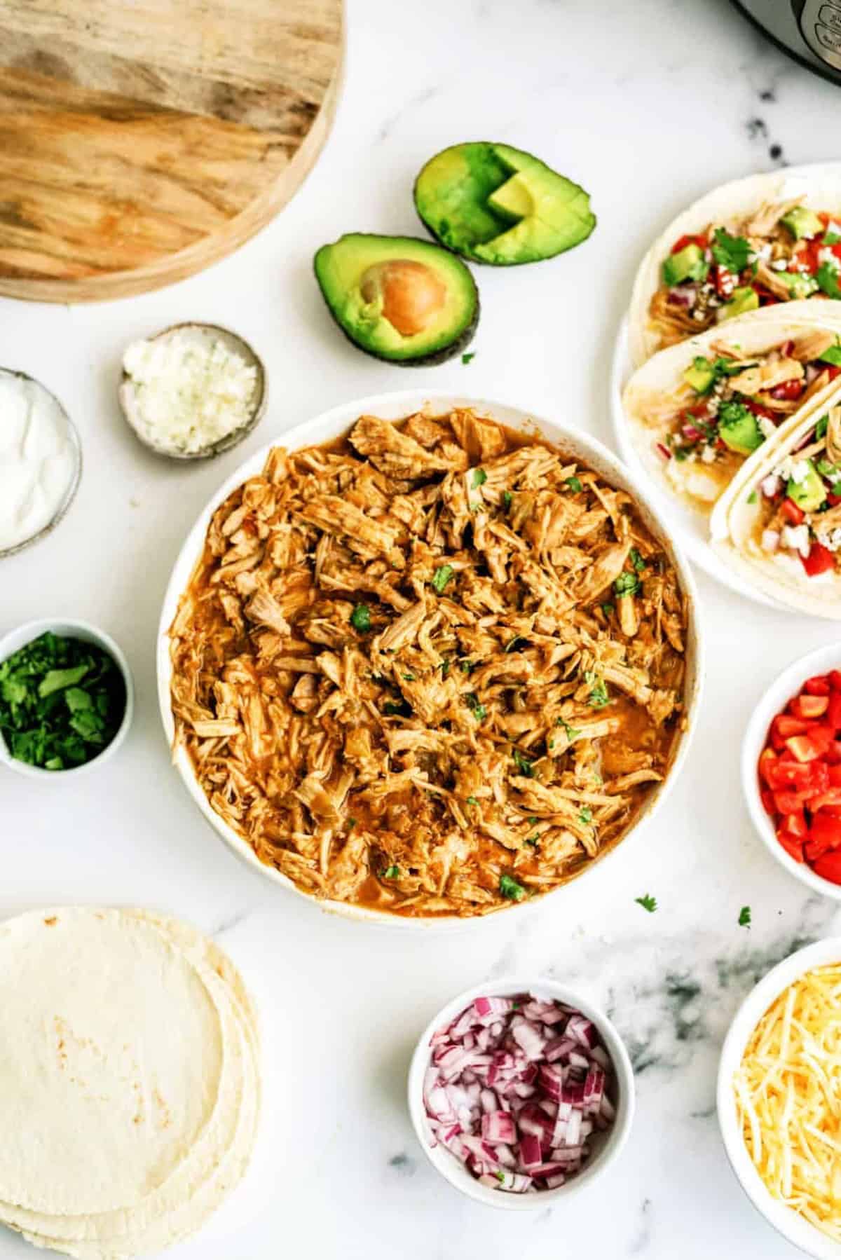 Shredded pork in a bowl surrounded by tortillas, diced red onion, chopped cilantro, avocado, tomatoes, cheese, sour cream, and assembled tacos on a white surface.