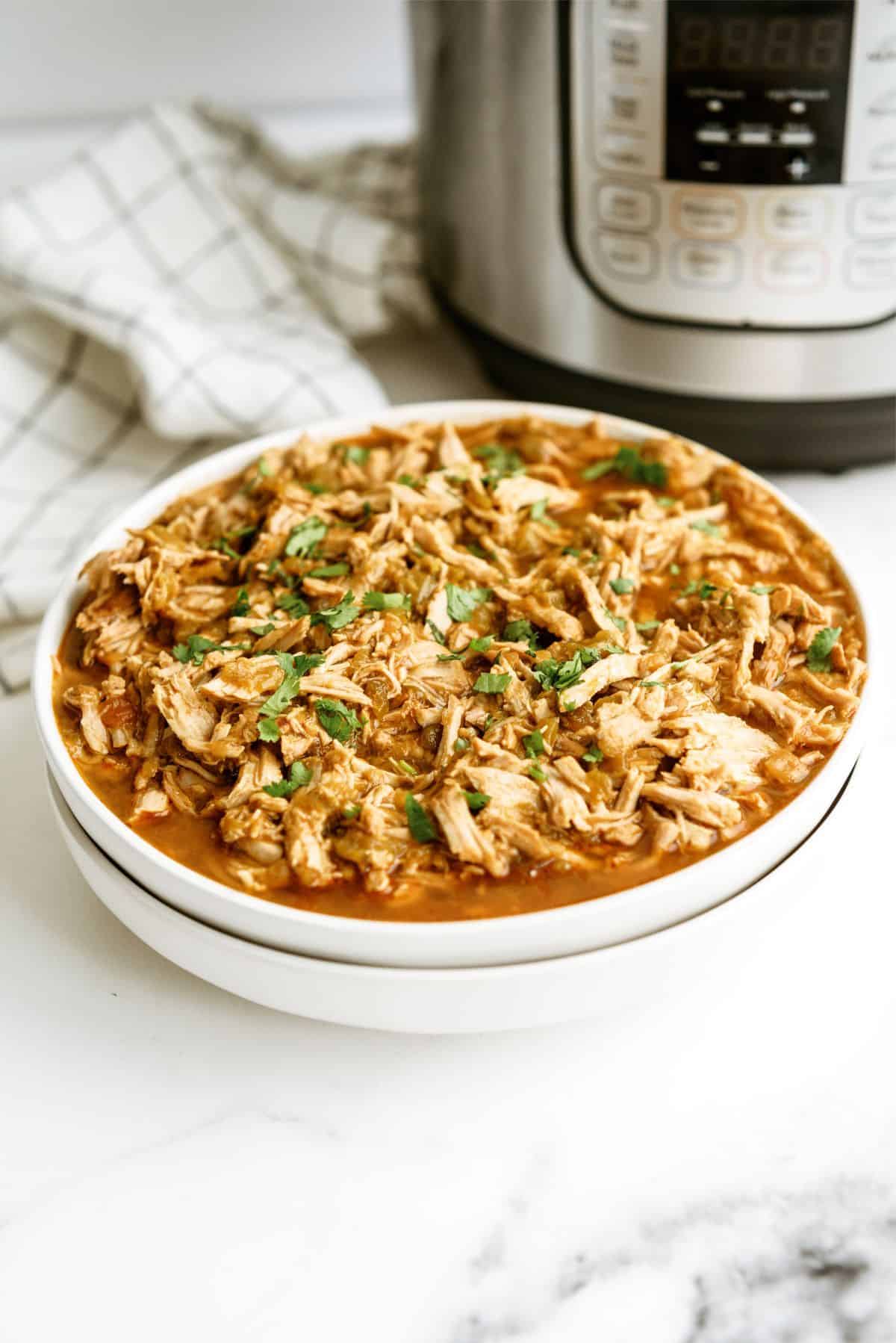 A white bowl filled with shredded pork in a red sauce, garnished with chopped herbs, sits on a counter next to an Instant Pot and a checkered cloth.