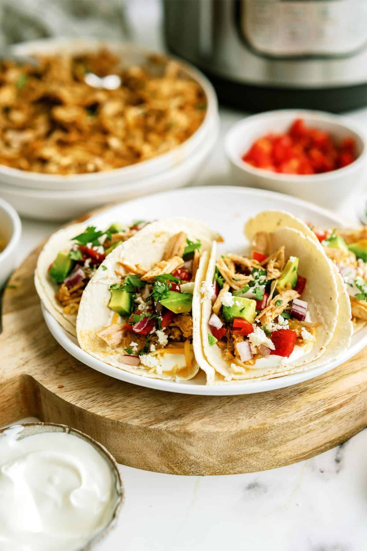 Three pork tacos with avocado, tomato, onion, and cilantro on a white plate, served with a side of sour cream; shredded chicken and diced tomatoes in bowls in the background.