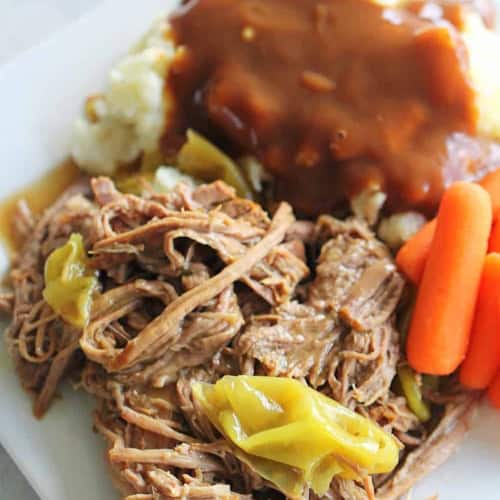 A plate with shredded beef and pepperoncini, mashed potatoes topped with brown gravy, and baby carrots.