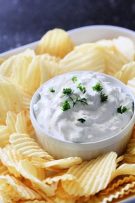 A bowl of creamy dip garnished with chopped herbs, surrounded by ridged potato chips.