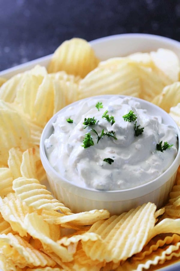 A bowl of creamy dip garnished with chopped herbs, surrounded by ridged potato chips.