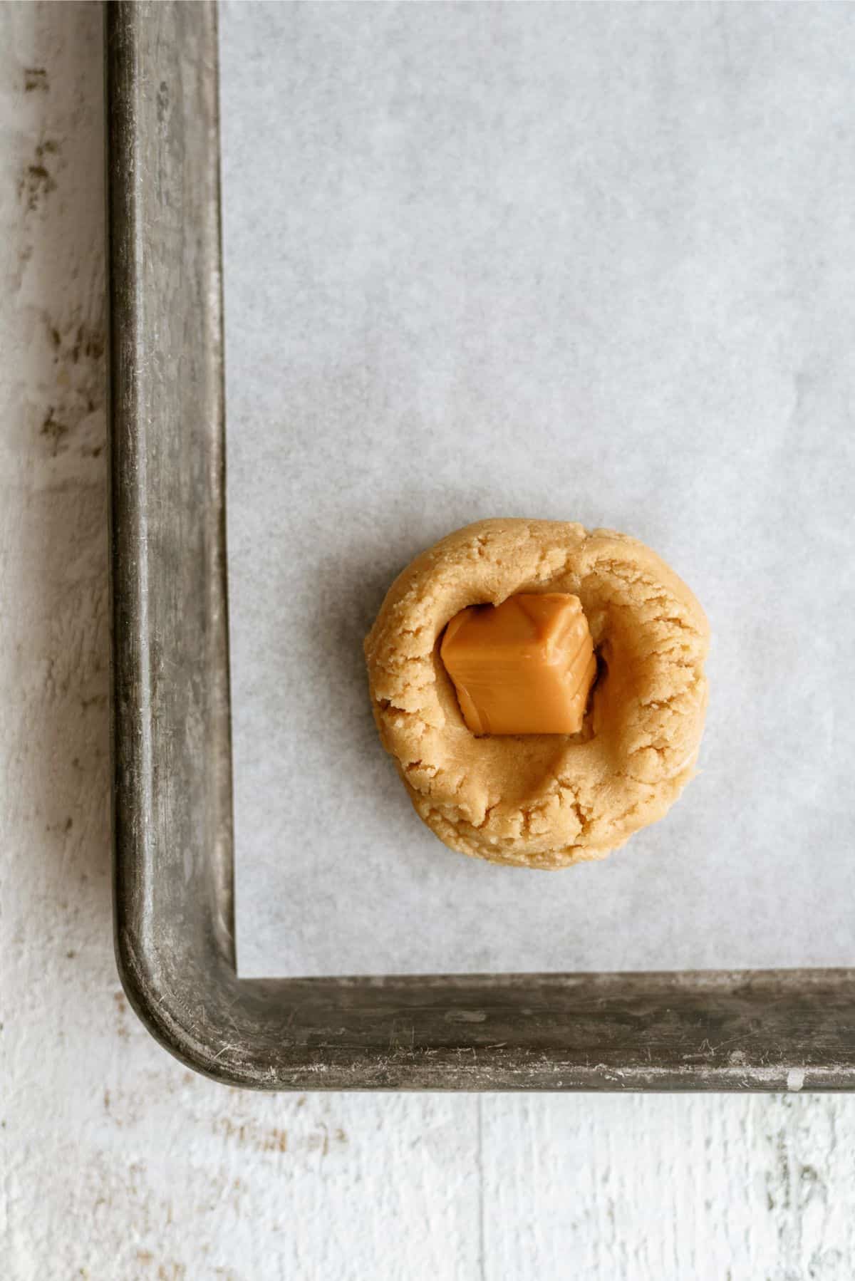 Flattened cookie ball with an unwrapped caramel in the middle on a baking sheet