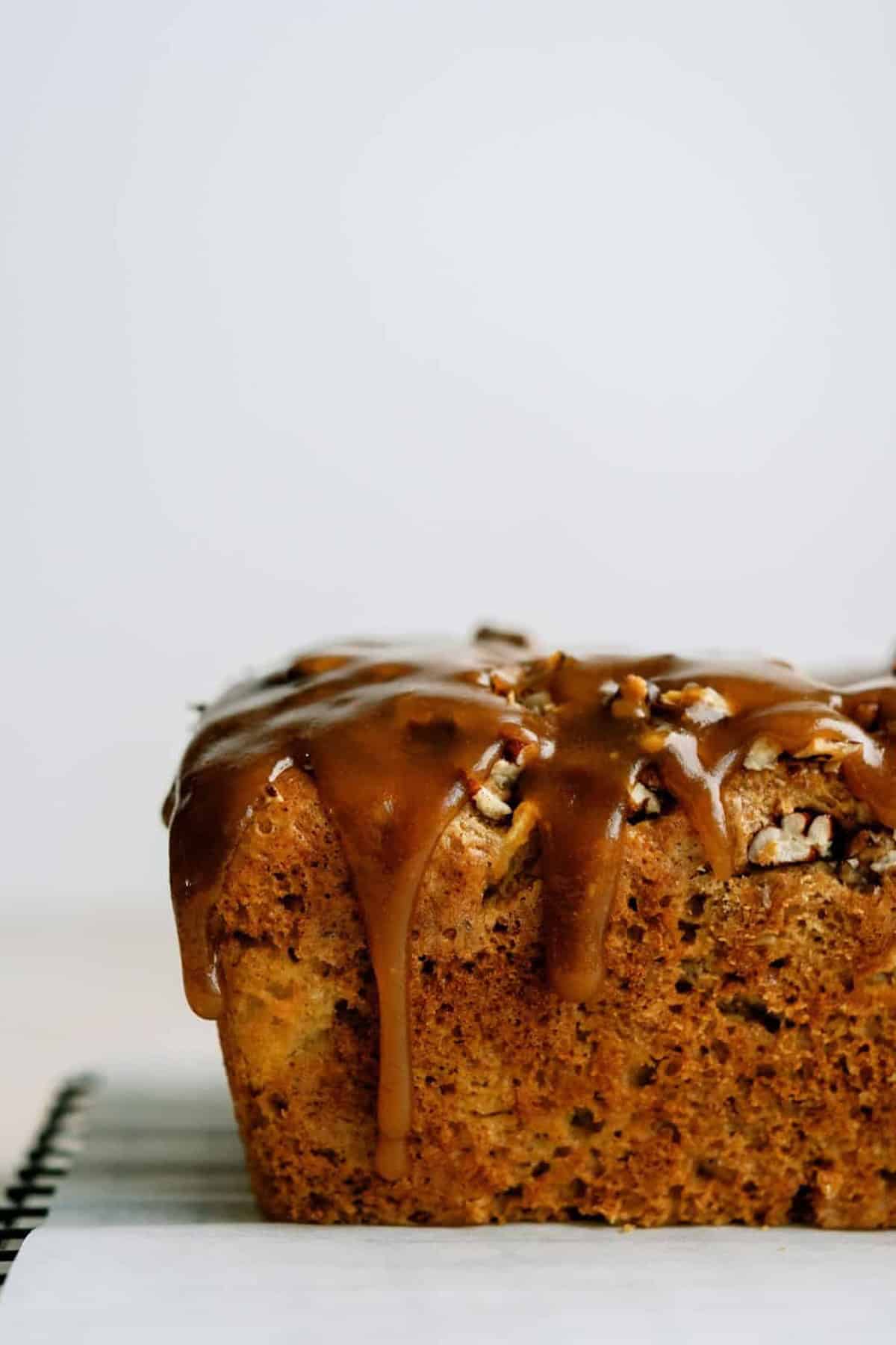 A close-up of a loaf of apple praline bread topped with a glossy caramel glaze and chopped nuts on a cooling rack.