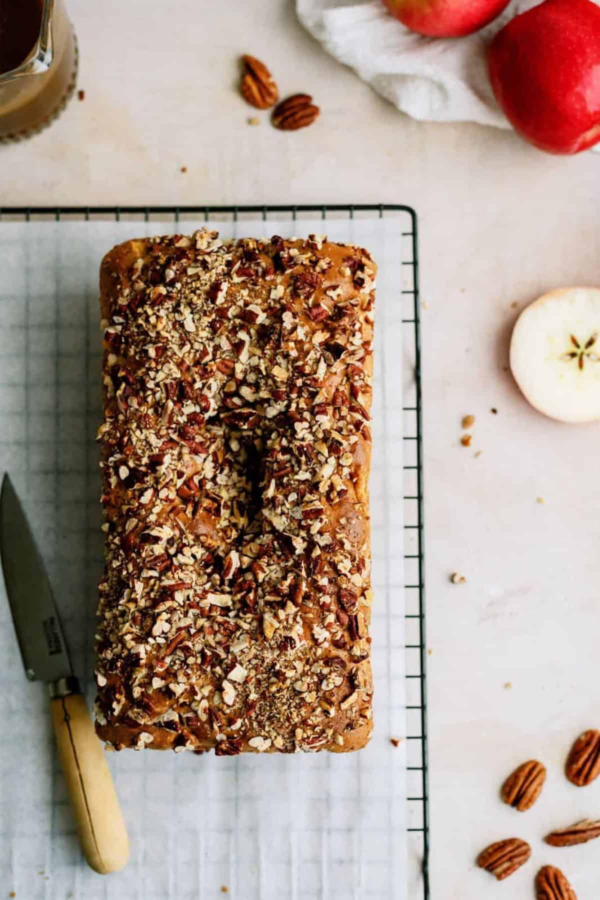 A loaf of bread topped with chopped nuts sits on a cooling rack beside a knife. Apples, pecans, and a white cloth are on the table.