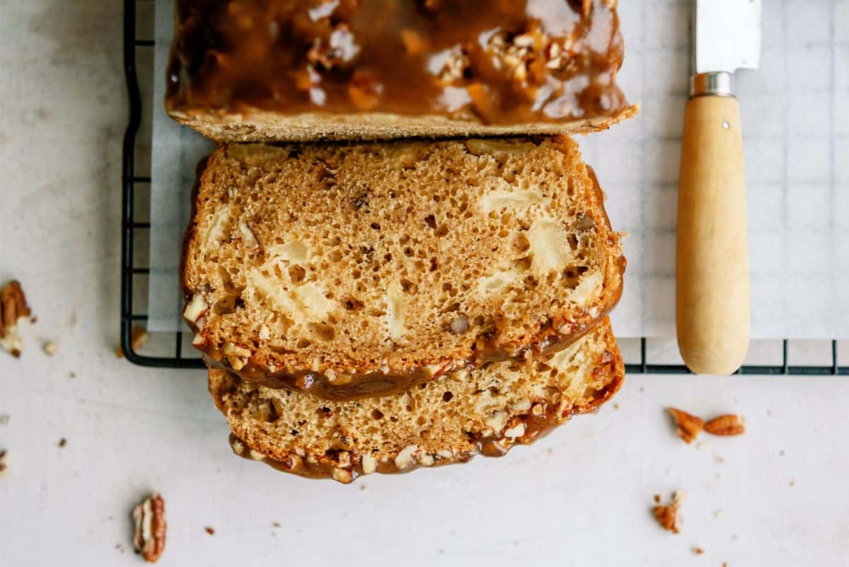 Sliced loaf of apple praline bread with visible apple chunks and pecans, displayed on a cooling rack next to a knife with a wooden handle.