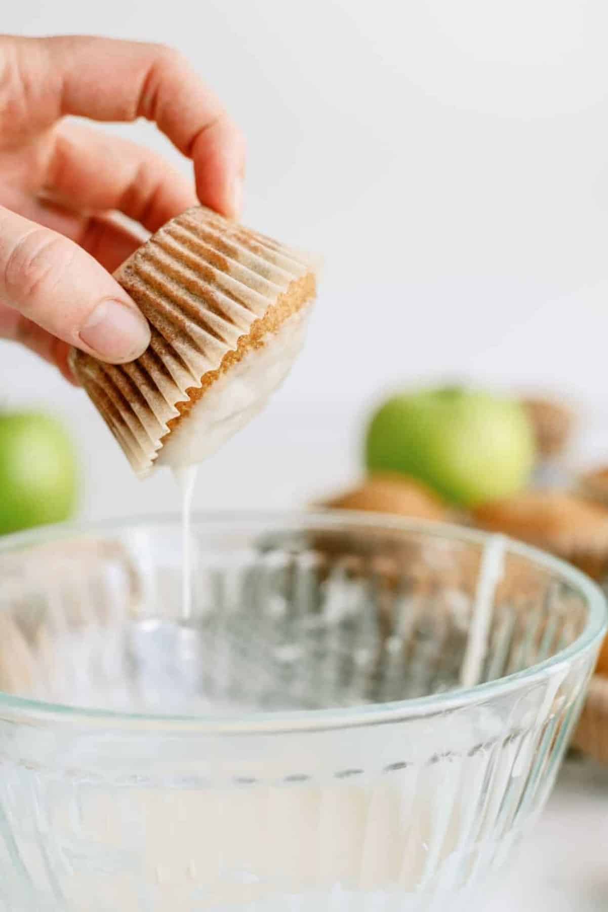 A hand dips a muffin into a bowl of glaze, with more muffins and green apples in the background.