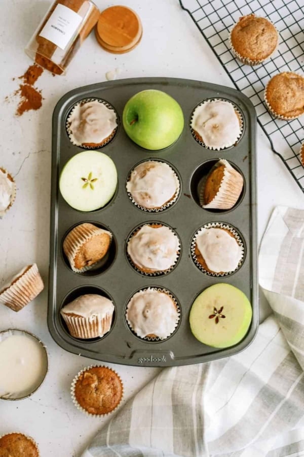 A muffin tin filled with apple muffins, some frosted and some plain. Green apple slices, a green apple, and a few muffins are scattered around. A spice jar and a cloth are present on the table.
