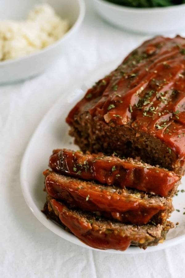 A meatloaf topped with a glaze, partially sliced, on a white plate. A bowl of mashed potatoes is visible in the background.