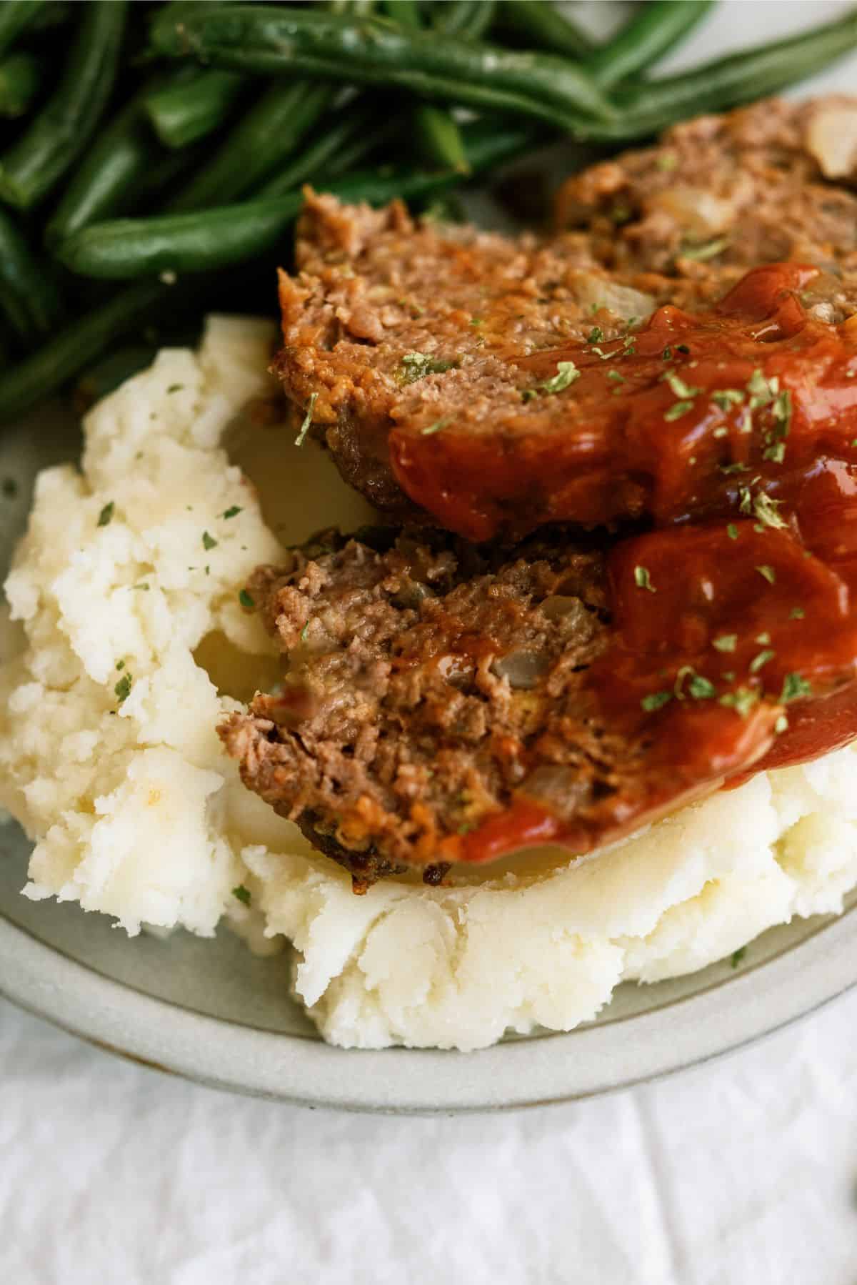 Top view of a plate with mashed potatoes topped with 2 slices of Copycat Cracker Barrel Meatloaf. With a side of green beans.