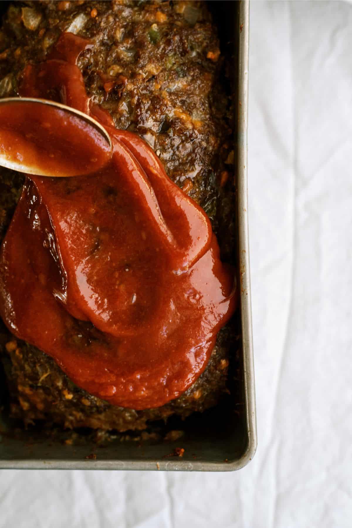 Spreading sauce with a spoon on top of meatloaf in a loaf pan.