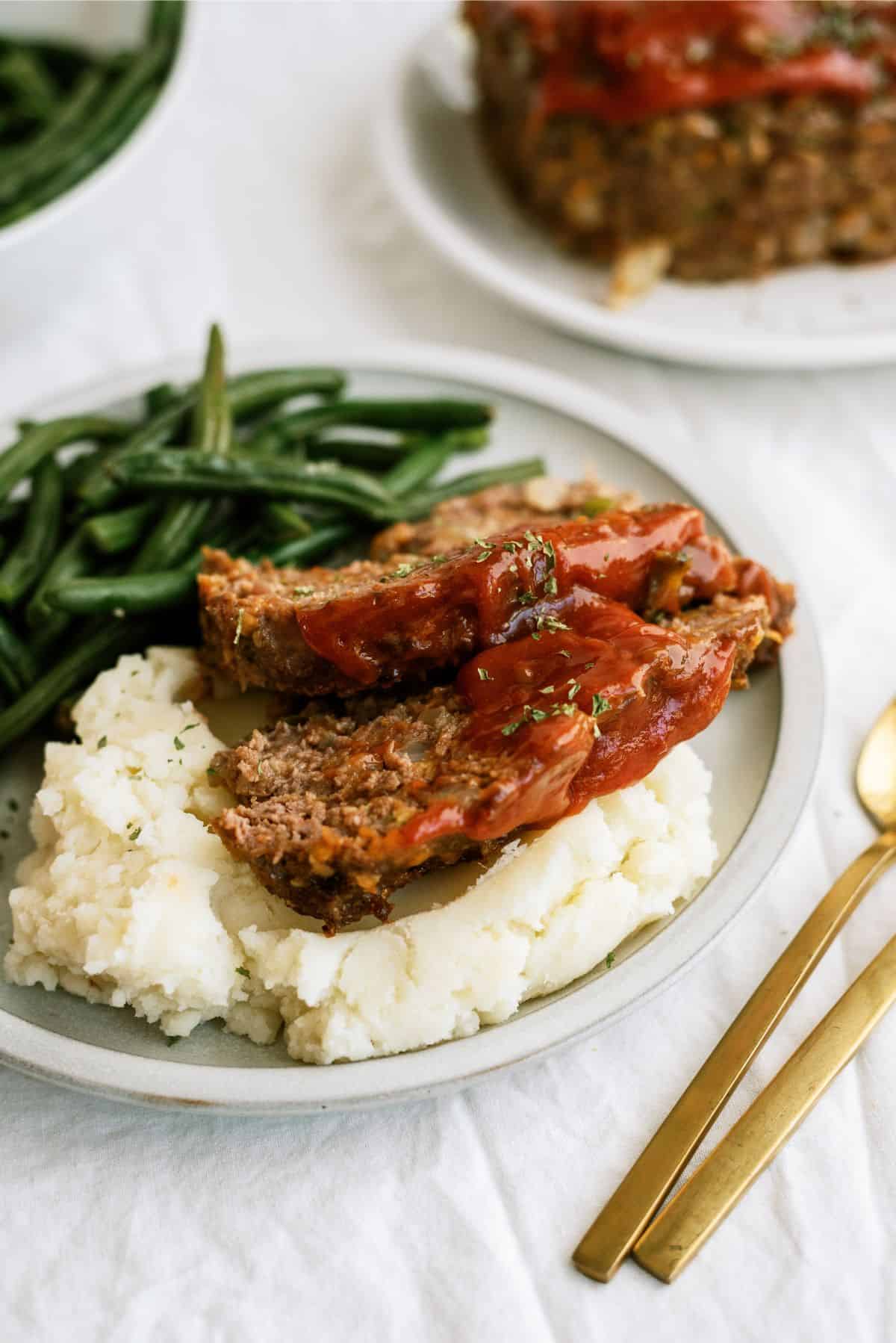 A plate with mashed potatoes and green beans. The massed potatoes are topped with 2 slices of Copycat Cracker Barrel Meatloaf.