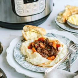A plate with mashed potatoes topped with beef stew and vegetables beside a piece of bread, with an Instant Pot in the background.