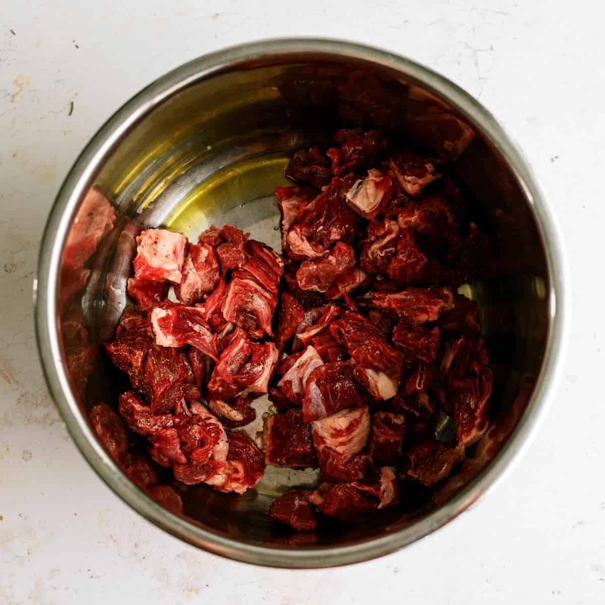 A stainless steel bowl containing raw chunks of red meat on a white surface.