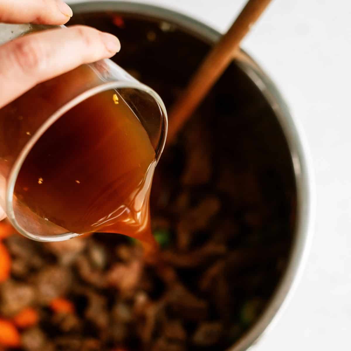 A hand holding a glass pouring brown liquid into a pot containing sliced vegetables and meat, with a wooden spoon visible inside the pot.