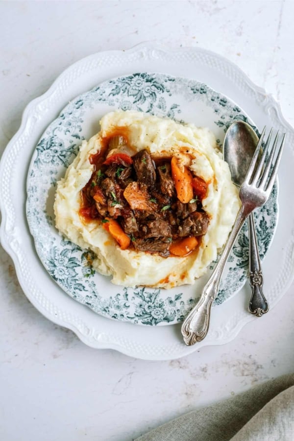 A plate with mashed potatoes topped with beef stew and carrots, served with a spoon and fork.