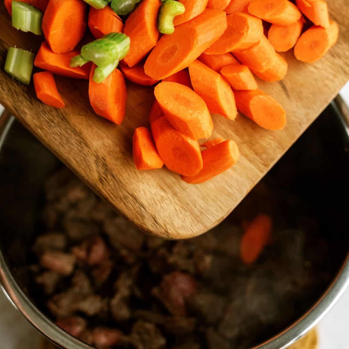 Chopped carrots and celery being added from a cutting board into a pot with cooked meat.