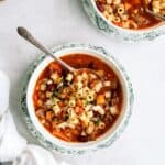 A bowl of minestrone soup with pasta, beans, and vegetables, placed on a table with a spoon inside. Another bowl with similar contents is partially visible in the background.