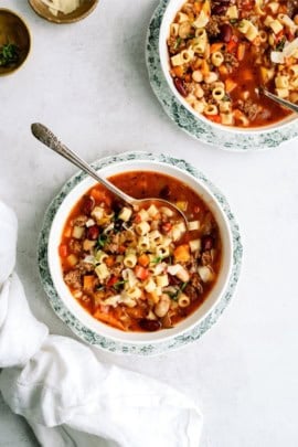 A bowl of minestrone soup with pasta, beans, and vegetables, placed on a table with a spoon inside. Another bowl with similar contents is partially visible in the background.