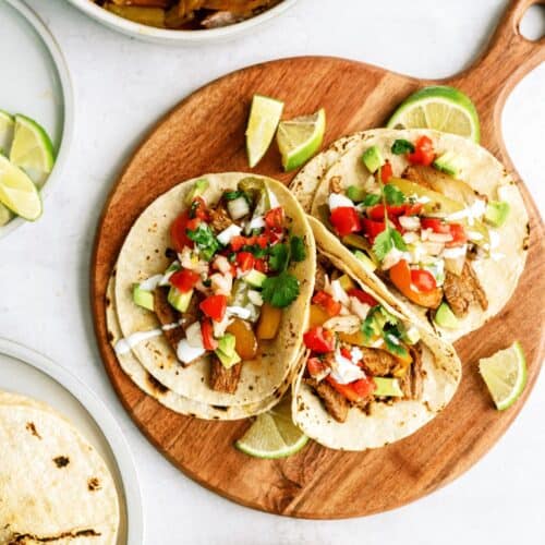 Three tacos filled with grilled meat, sautéed peppers, onions, tomatoes, cilantro, and diced onions on a wooden board, with lime wedges and salsa nearby.