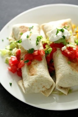 Two burritos topped with sour cream and chopped cilantro, served with diced tomatoes and shredded lettuce on a white plate.