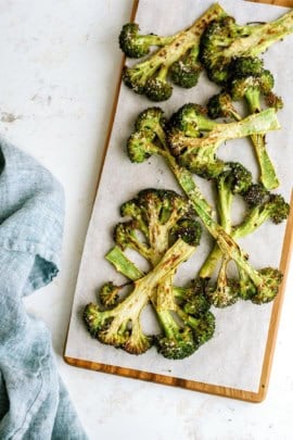 Roasted broccoli slices on a parchment-lined wooden board, with a gray cloth napkin to the left.