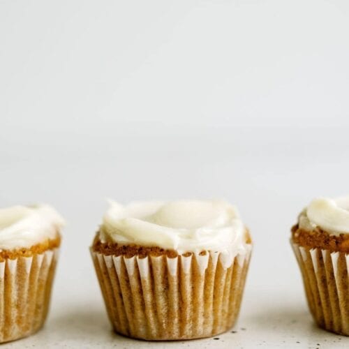 Three frosted muffins in white liners are evenly spaced in a row against a plain white background.