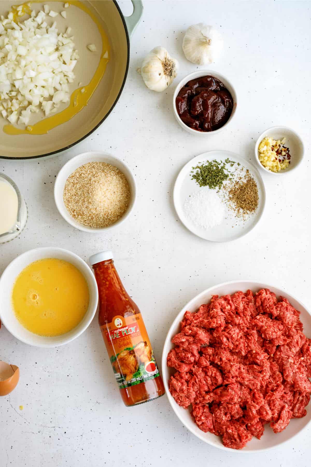 Ingredients on a countertop: chopped onions in a pan, raw ground beef, eggs, breadcrumbs, spices, minced garlic, tomato paste, garlic bulbs, and a bottle of spicy sauce.