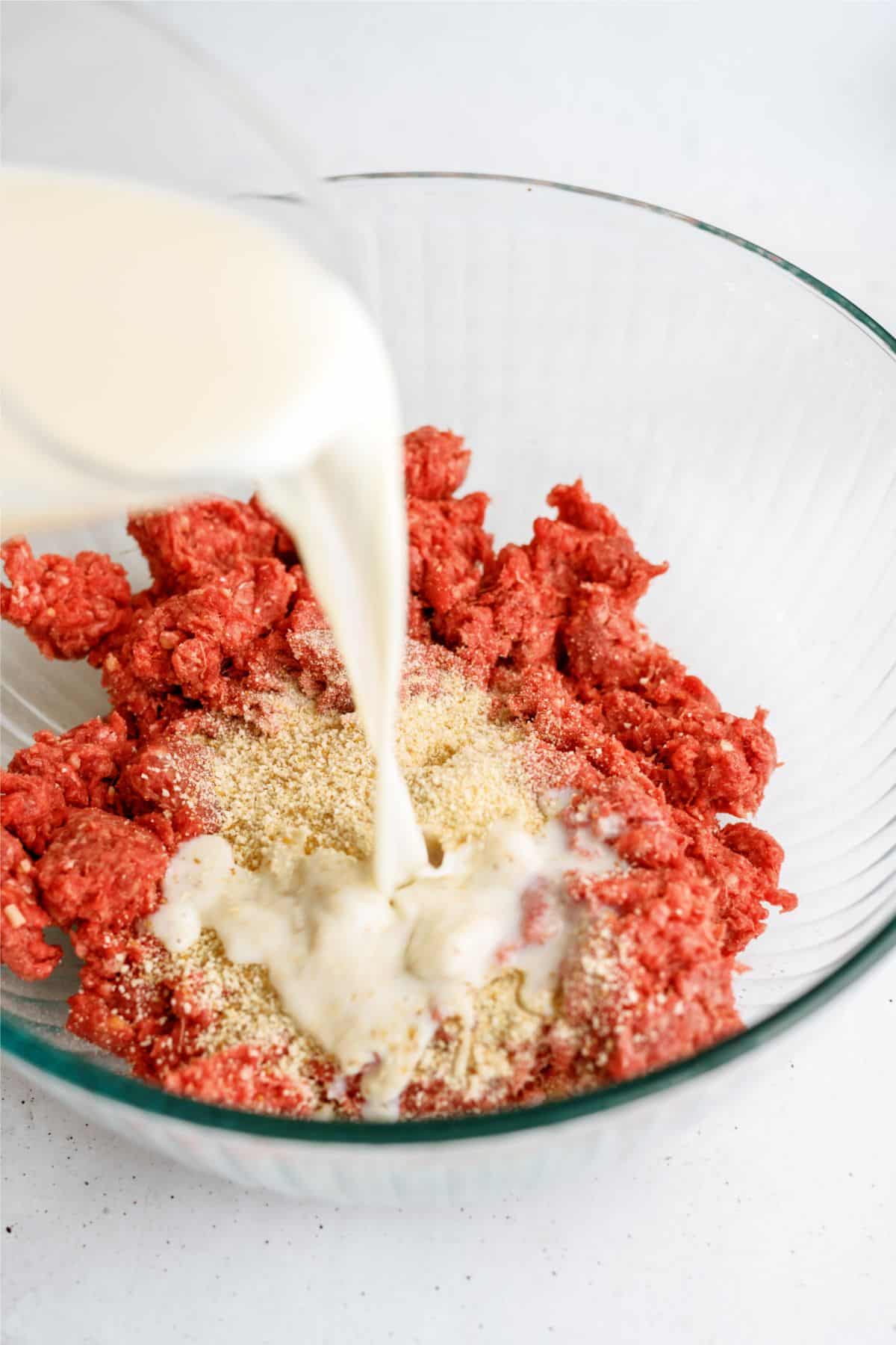 Pouring milk into a glass bowl containing ground meat and breadcrumbs.