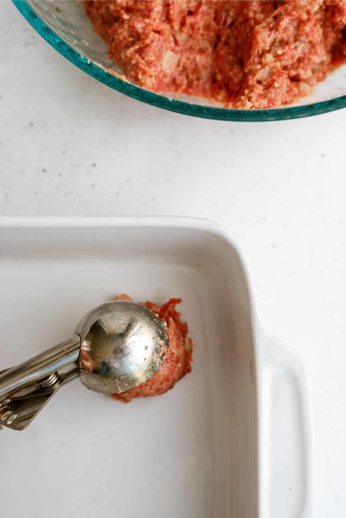 Metal scoop placing ground meat into a white baking dish, with a bowl of more ground meat in the background.