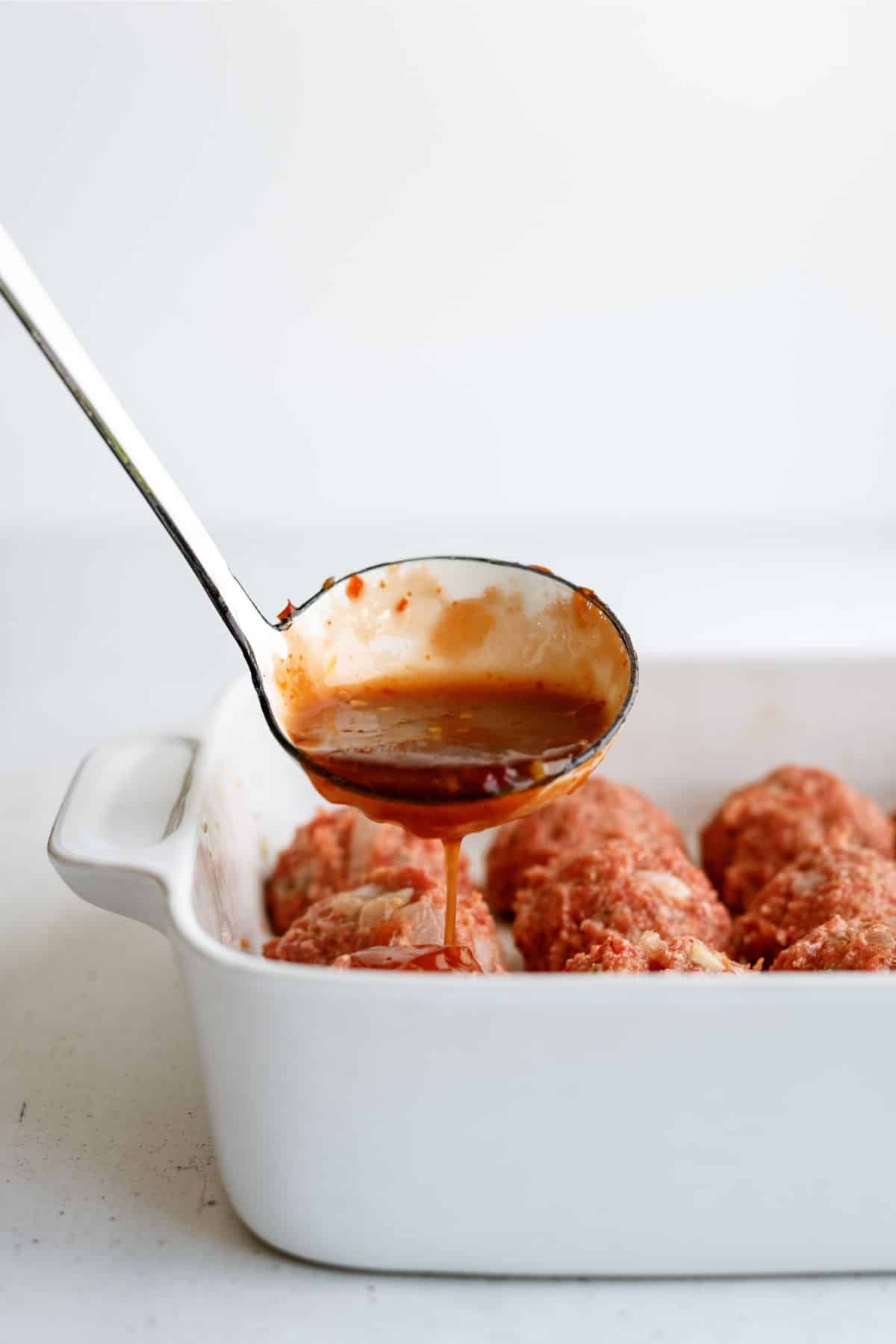 A ladle pouring sauce over a row of meatballs in a white baking dish.