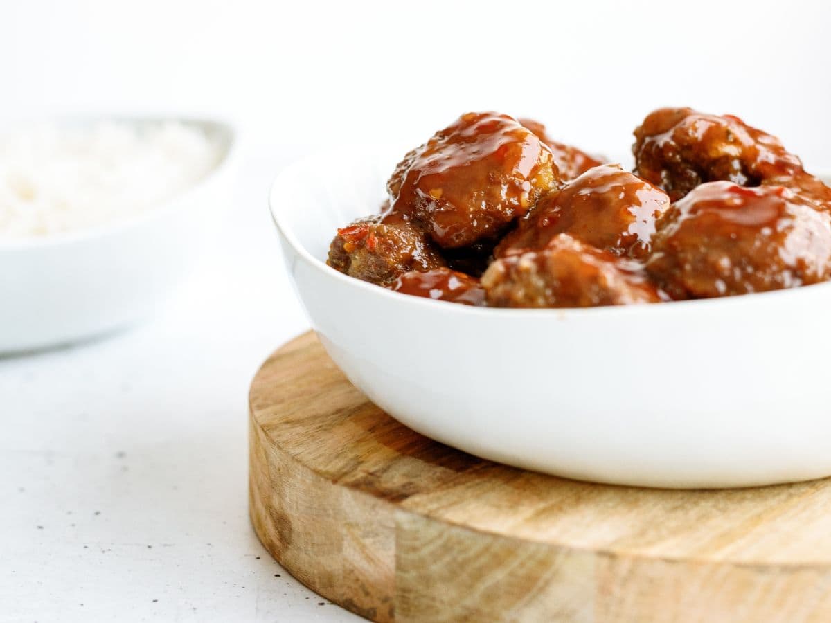 A white bowl of glazed meatballs is placed on a wooden board. A separate bowl of white rice is in the background.