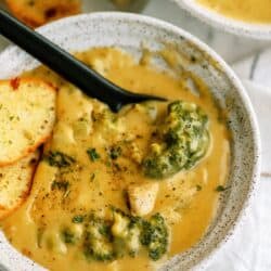 A bowl of creamy broccoli cheddar soup garnished with herbs, served with two pieces of toasted bread and a black spoon on a light-colored surface.