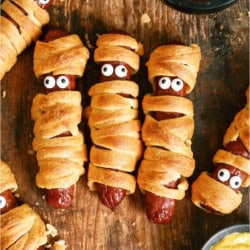 Halloween Mummy Dogs lined up on a wooden background with a small bowl of ketchup and mustard.