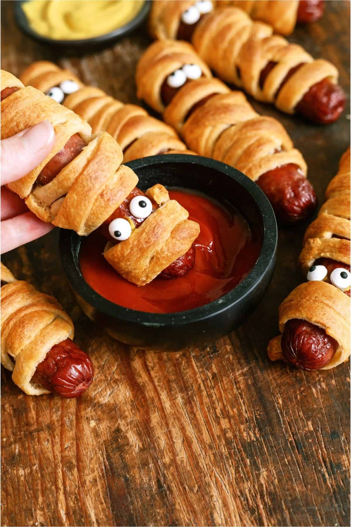 A hand dipping a Halloween Mummy Dog into a small cup of ketchup.
