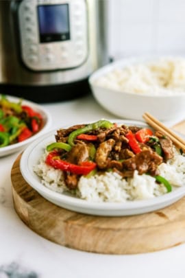 A plate of beef stir-fry with bell peppers on white rice, garnished with chopsticks, with a bowl of rice and vegetables in the background.