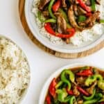 Two plates of stir-fried beef with red and green bell peppers served over white rice, with an additional bowl of plain white rice beside them.