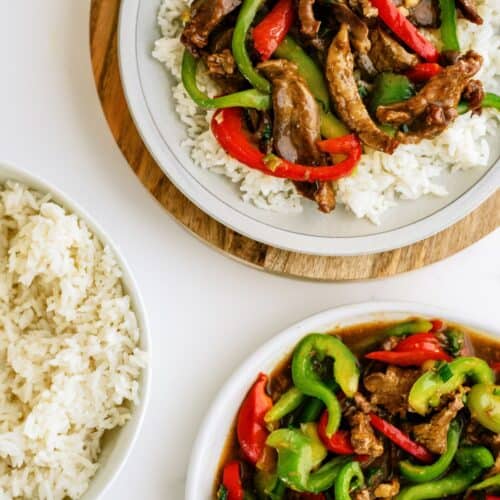 Two plates of stir-fried beef with red and green bell peppers served over white rice, with an additional bowl of plain white rice beside them.