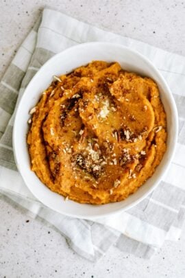 A white bowl filled with mashed sweet potatoes, topped with brown sugar, cinnamon, and chopped nuts, placed on a gray and white striped cloth.