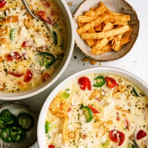 Two bowls of creamy soup topped with sliced jalapeños, tomatoes, crumbled cheese, and tortilla strips, with extra jalapeños and tortilla strips in small bowls nearby.