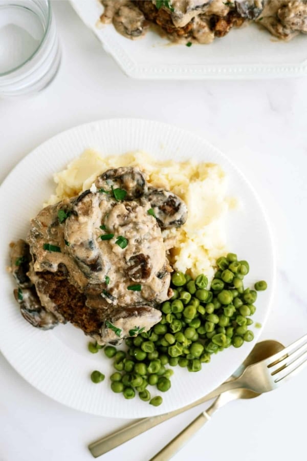 A plate of mashed potatoes, peas, and a creamy mushroom dish. A fork and knife are beside the plate.