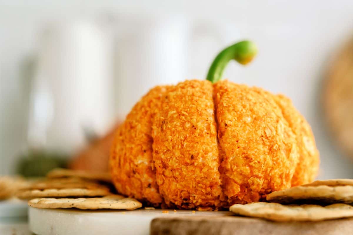 A cheese ball shaped like a pumpkin with a green stem on top, surrounded by crackers on a marble surface.