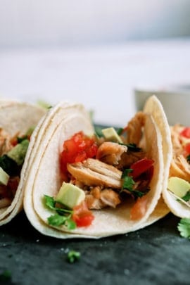 Two tacos filled with shredded meat, diced tomatoes, avocado, and cilantro are arranged on a plate with a bowl visible in the background.